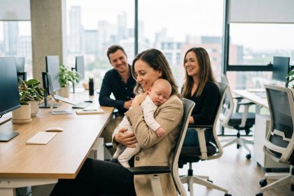 Femme professionnelle tenant un bébé dans un bureau moderne, entourée de collègues souriants, illustrant le lien humain au travail